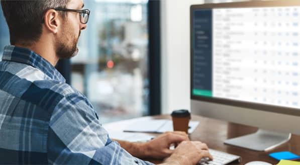 Man in a blue and white plaid shirt working on his desktop computer at his desk